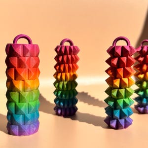 May include: Four colorful, stacked, geometric sculptures in a rainbow of hues. Each sculpture has a handle at the top. The colors transition from purple to blue, green, yellow, orange, and red. The background is a solid, light peach color.
