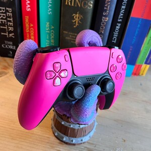 Peut inclure: Une manette rose de PlayStation 5 repose sur un présentoir décoratif. La manette a des boutons noirs et une face rose. Le présentoir est marron et gris, avec un design en forme de tentacule. Des livres sont en arrière-plan.