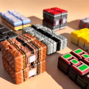 Könnte beinhalten: Eine Auswahl an farbenfrohen, geometrischen Puzzle-Würfeln. Die Würfel weisen verschiedene Farbschemata auf, darunter Orange mit schwarzen Streifen, Rot und Grau, Blau und Gelb sowie Schwarz und Weiß. Die Würfel sind auf einer hellen Oberfläche angeordnet.