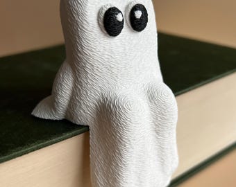 May include: A white ghost-shaped bookend with black eyes, perched on a green book. The ghost has a textured surface, giving it a unique, handmade appearance. The bookend adds a touch of whimsy to any bookshelf.