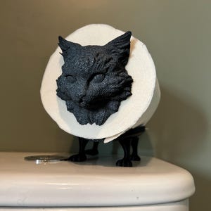 Op de afbeelding: Een zwarte katvormige toiletpapierhouder. De kop van de kat is gedetailleerd gebeeldhouwd, met de toiletrol die een halo rond zijn gezicht vormt. De houder is op een toilet gemonteerd.
