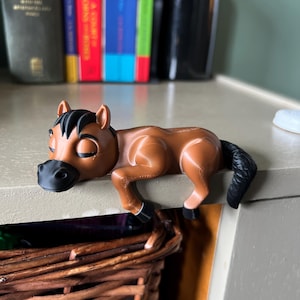 May include: A brown plastic horse figurine with a black mane, tail, and hooves. The toy horse is lying down with its head resting on a surface. The horse is a decorative item.