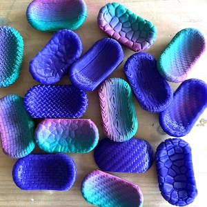 May include: A selection of small, oval soap dishes in diverse colours and textures. The dishes showcase a variety of designs, including woven, brick, and wood grain patterns. The colour scheme includes shades of blue, purple, teal, and pink.