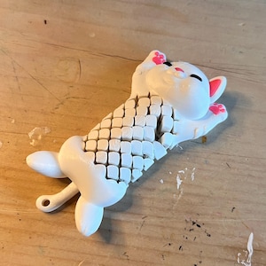 Puede incluir: Figura de gato blanco impresa en 3D con un diseño segmentado en la espalda. El gato tiene detalles en rosa en las orejas y las patas, y está tumbado. La figura está sobre una superficie de madera.