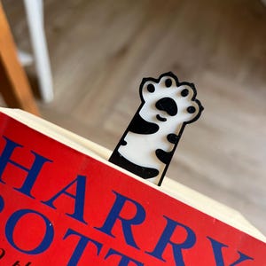 May include: A black and white cat paw bookmark is inserted into a book with a red cover. The book's title, "HARRY POTTER and the Goblet of Fire" is visible in blue lettering. The bookmark is made of a glossy material.