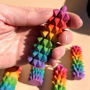 May include: A selection of vibrant, rainbow-coloured, geometric objects. Each object has a spiky, vertical design, with each section displaying a different colour of the rainbow. The objects are held in a hand.