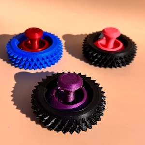 May include: Three colorful 3D-printed tops. One is blue with a red top, another is black with a pink top, and the third is black with a purple top. The tops have a gear-like design around the base.