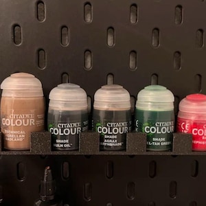 Puede incluir: Cinco pequeños frascos de pintura Citadel Colour, organizados en una placa perforada negra. Los frascos están etiquetados con el tipo de pintura, el tono y el nombre. Los colores de la pintura son marrón, negro, gris, verde y rojo.