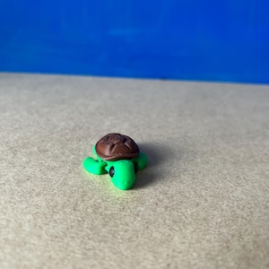 May include: A small green turtle figurine with a brown shell. The turtle has black eyes and a simple design.