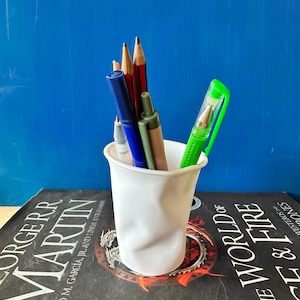 May include: A white ceramic pencil holder with a crumpled texture, holding a variety of pens and pencils, including a green pen, a blue pen, a grey pen, a red pencil, and a grey pencil. The holder is sitting on a book with the text "George R.R. Martin" and "A World of Ice and Fire" visible.