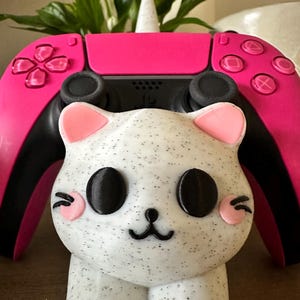 Pode incluir: Um suporte para controlador em forma de gato branco com olhos pretos e bochechas cor-de-rosa. O suporte está segurando um controlador PlayStation 5 rosa e preto.