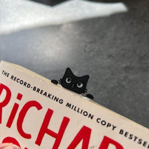 May include: A black cat-shaped bookmark made of a flat material, featuring a grumpy facial expression with white eyes and small paws extending to the sides. The bookmark is inserted into the top edge of a book with the text THE RECORD-BREAKING MILLION COPY BESTSELLER visible on the cover.