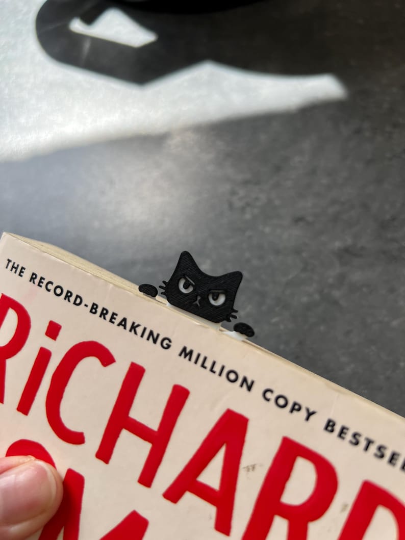 May include: A black cat-shaped bookmark made of a flat material, featuring a grumpy facial expression with white eyes and small paws extending to the sides. The bookmark is inserted into the top edge of a book with the text THE RECORD-BREAKING MILLION COPY BESTSELLER visible on the cover.