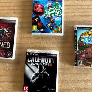 Puede incluir: Una colección de carcasas de juegos de PS3 en miniatura. Los títulos incluyen "LittleBigPlanet 2", "Shadows of the Damned" y "Call of Duty: Black Ops II". Las carcasas de los juegos presentan ilustraciones coloridas y títulos de juegos.