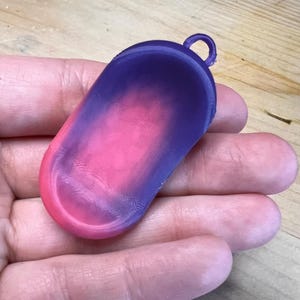 May include: A 3D-printed, oblong pendant with a colour gradient. The top is a deep purple, transitioning to pink in the centre, and fading to a lighter pink at the bottom. It has a small loop at the top for attachment.