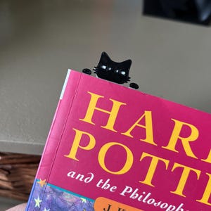 May include: A black cat-shaped bookmark peeking over the top edge of a book titled Harry Potter and the Philosopher's Stone by J.K. Rowling. The bookmark features a stylized feline head with white eyes and small paws resting on the book cover.