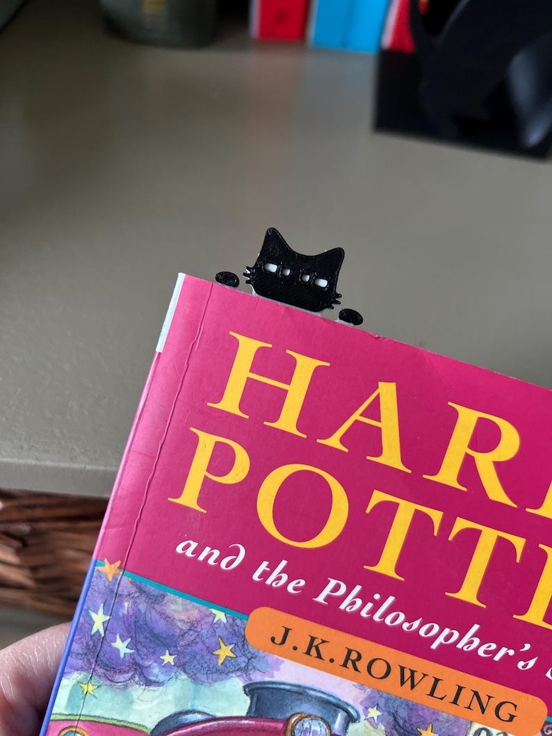 May include: A black cat-shaped bookmark peeking over the top edge of a book titled Harry Potter and the Philosopher's Stone by J.K. Rowling. The bookmark features a stylized feline head with white eyes and small paws resting on the book cover.