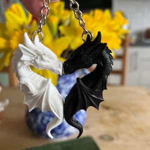 May include: A keyring with two dragon pendants, one white and one black, arranged in a heart shape. The dragons have detailed wings and scales. The keyrings are connected by a silver chain.