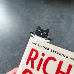 May include: A black and white cat-shaped bookmark made of flat material, featuring a smiling face and two small paws extending outward. The bookmark is positioned at the top edge of a book with white pages and large red text on the cover.