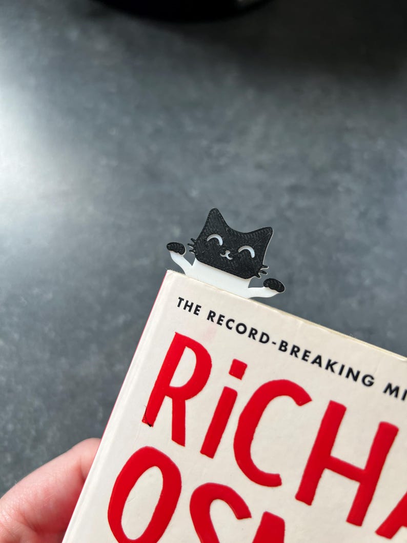 May include: A black and white cat-shaped bookmark made of flat material, featuring a smiling face and two small paws extending outward. The bookmark is positioned at the top edge of a book with white pages and large red text on the cover.