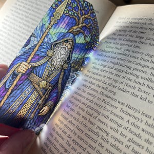 May include: A bookmark featuring a stained-glass style illustration of a bearded figure with an eyepatch, wearing a blue cloak and holding a tall staff. The design includes a large tree and a castle in the background with vibrant purple, blue, and gold tones.