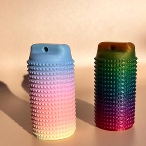 Könnte beinhalten: Zwei strukturierte Kerzenhalter in Zylinderform. Einer hat einen Farbverlauf von Blau, Rosa und Gelb, der andere von Grün, Lila und Rot. Beide haben oben ein kleines Loch.