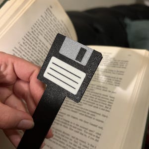 Könnte beinhalten: Schwarzes Lesezeichen in Form einer Diskette mit einem weißen Etikett und einem schwarzen Griff. Das Lesezeichen liegt auf einem Buch.