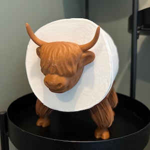 May include: A brown Highland cow-shaped toilet paper holder with a white roll of toilet paper. The holder is made of plastic and has a realistic design.
