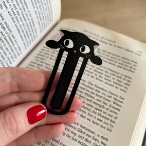 Puede incluir: Marcapáginas de gato negro con un diseño caprichoso. El marcapáginas presenta una silueta de gato negro con ojos grandes y expresivos y detalles en rosa en las orejas. Está hecho de un material duradero y está diseñado para marcar una página en un libro.