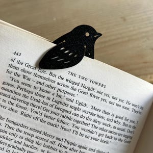 Puede incluir: Un marcapáginas negro con forma de pájaro, hecho de un material oscuro, que descansa sobre las páginas de un libro. El marcapáginas tiene un diseño sencillo y estilizado con detalles recortados. El texto del libro es visible.