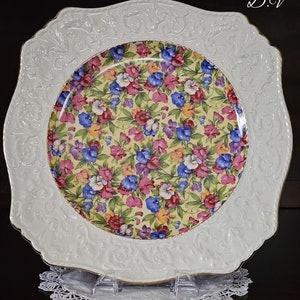 May include: A white ceramic serving plate with a raised, ornate border and a floral pattern in shades of pink, blue, yellow, and green. The plate has a gold rim and the initials "D.V." are visible in the upper right corner.