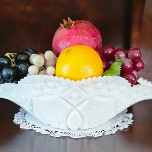 May include: A white milk glass bowl with a scalloped edge and a raised design, filled with a variety of fruit, including grapes, a pomegranate, and an orange.