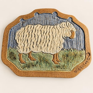 May include: A rectangular ceramic plaque with rounded corners featuring a relief of a white sheep standing on green grass against a blue sky. The border is a speckled tan clay. The artist signature J. Peters and a copyright symbol are visible.