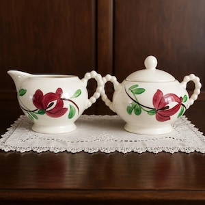 May include: A cream-colored ceramic creamer and a lidded sugar bowl set, both featuring hand-painted dark pink floral motifs with green leaves and distinctive beaded handles. The pieces have a smooth, glossy finish and a rounded, pedestal base.