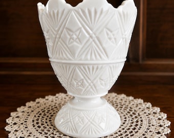May include: A white milk glass vase featuring a scalloped rim and a pressed diamond and starburst pattern. The vessel has a wide, flared opening that tapers down to a pedestal base, with the intricate geometric design covering the entire exterior.