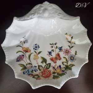 May include: A white ceramic serving dish shaped like a seashell with a raised design. The dish features a floral pattern with a butterfly in the center. The flowers are in shades of pink, blue, yellow, and red.