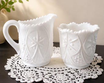 Op de afbeelding: Een witte melkkan en een bijpassende suikerpot van melkglas, beide voorzien van een geperst glaspatroon met sterren, geometrische lijnen en kralenranden. De melkkan heeft een gebogen handvat en een schenktuit, de suikerpot is open.