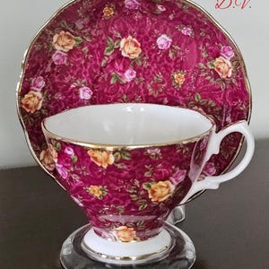 May include: A vintage teacup and saucer set. The set has a deep burgundy background with a floral pattern of yellow and pink roses. The cup and saucer have gold trim and a white interior.