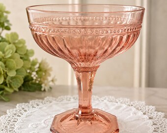 May include: A light pink glass dessert bowl featuring a wide, shallow basin with vertical fluted detailing and a beaded rim. The bowl is supported by a faceted stem and a sturdy, hexagonal base, crafted from translucent, rose-tinted pressed glass.