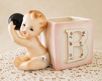 May include: A ceramic figurine of a baby sitting next to a square block with the letter B. The baby has light skin, brown hair, and is holding a black ball. The block is pale pink with a white letter B decorated with small red and green flowers.