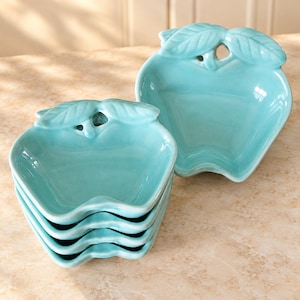 May include: A set of five glossy, light turquoise ceramic bowls shaped like apples. Each bowl features a sculpted stem and two leaves at the top edge. The bowls are stacked together in a vertical pile, with one bowl positioned separately to the right.