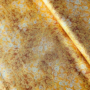 Op de afbeelding: Close-up van een stof met een herhaald patroon van gouden, bruine en lichtblauwe wervelingen. De stof heeft een getextureerde uitstraling en een subtiele glans. Het ontwerp doet denken aan een abstract schilderij.