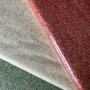 May include: Close-up of three rolls of wrapping paper. One is red with gold swirls, another is white with gold swirls, and the third is green with gold swirls. The paper has a glossy finish.