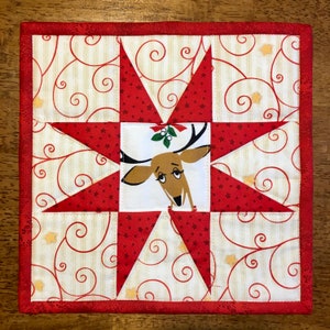 Peut inclure: Un carré matelassé rouge et blanc avec un motif d'étoile. Le centre de l'étoile présente un renne brun avec une branche de houx sur ses bois. L'arrière-plan est de couleur crème avec un motif de spirales rouge et blanc.