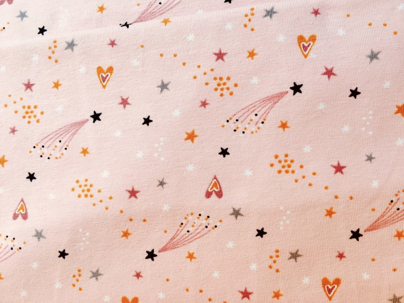 May include: Pink fabric with a repeating pattern of stars, hearts, and shooting stars. The stars are in shades of black, gray, and orange. The hearts are orange and pink. The shooting stars are black with orange and pink trails.