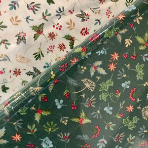 Op de afbeelding: Twee rollen stof met een feestelijk vakantiepatroon. Eén rol is crèmekleurig met hulst, bessen en stervormige bloemen. De andere rol is donkergroen met hetzelfde patroon. De stof is waarschijnlijk voor knutselen of seizoensdecoratie.