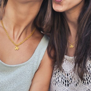 May include: Two women are taking a selfie. The woman on the left is wearing a light blue tank top and a gold necklace with a puzzle piece pendant. She is wearing tortoise shell sunglasses. The woman on the right is wearing a white lace tank top and a gold necklace with a puzzle piece pendant. She is wearing brown sunglasses.