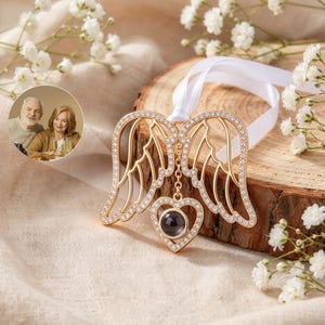 May include: A gold-toned metal ornament featuring two symmetrical angel wings encrusted with small clear crystals. A central heart-shaped pendant hangs from the wings, containing a circular dark lens. A white ribbon is attached to the top.