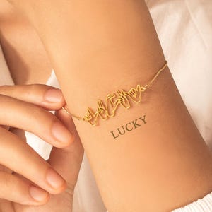 May include: A gold-colored bracelet spelling out the word "HAPPY" in sign language. The bracelet is worn on a wrist with a "LUCKY" tattoo. The background is a white shirt.