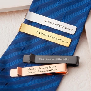 May include: Four tie clips with engraved text. The tie clips are silver, gold, black, and rose gold. The text on the tie clips reads "Father of the Bride", "Father of the Groom", "September 13th, 2024", and "Thank you for raising the man I am so lucky to marry. Katie".